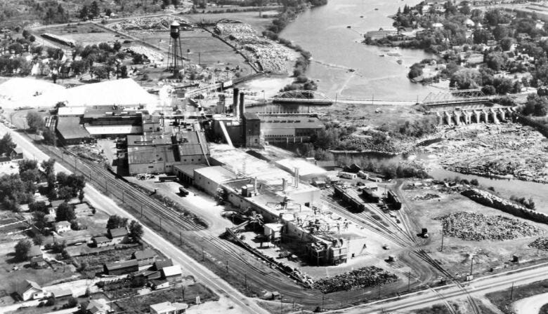 Black and white aerial photo showing an industrial paper mill beside a river, surrounded by homes and residential streets.