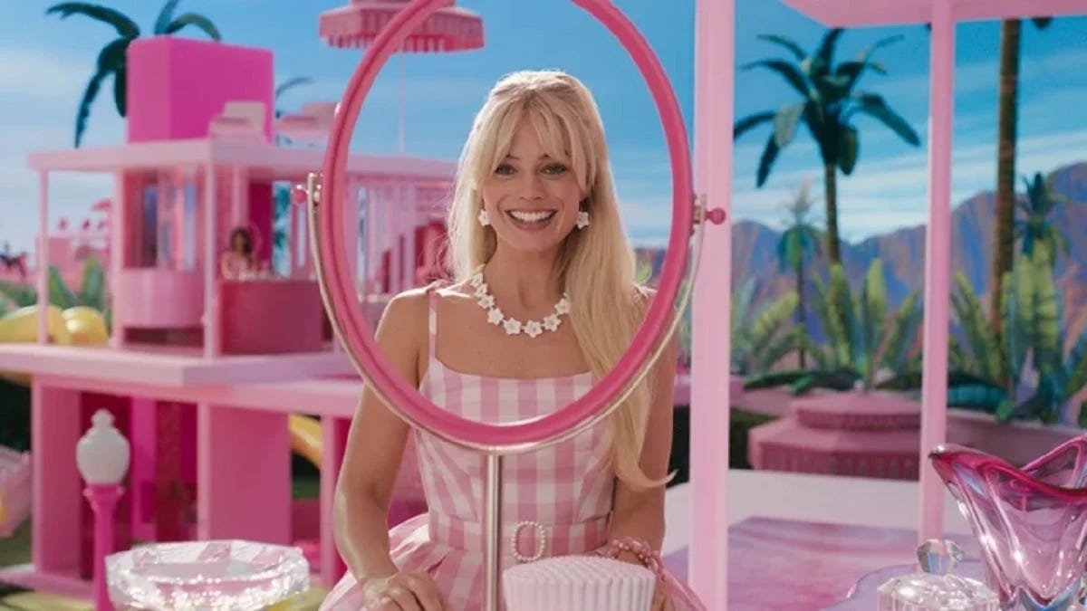 Scena del film Barbie in cui Margot Robbie, bionda e vestita di rosa, guarda attraverso la cornice (rosa) di un finto specchio. Sullo sfondo la casa di Barbie (rosa again), con palme e un cielo azzurro