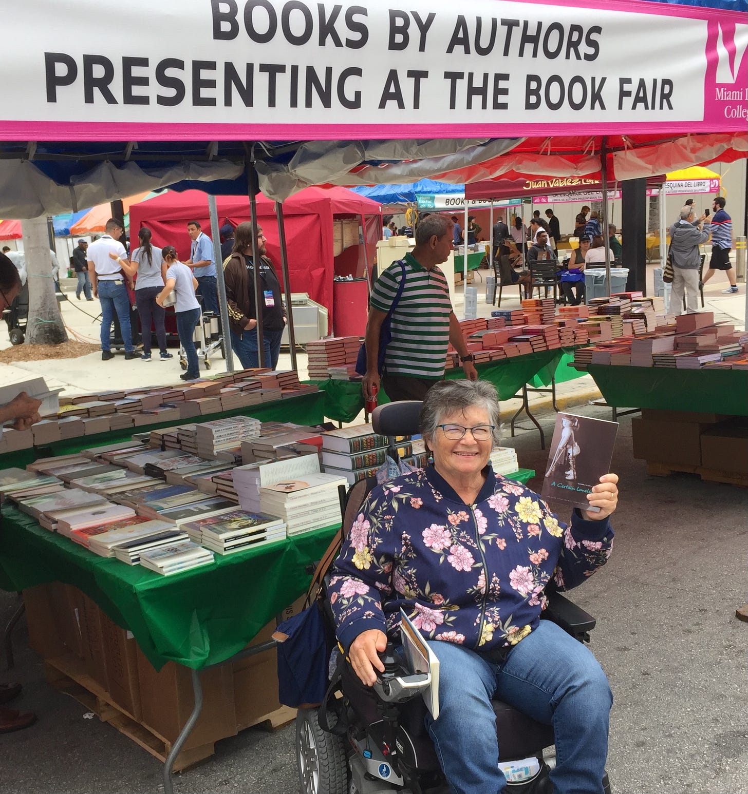 A white woman in a flowered jacked seated in her power chair holding up a copy of her memoir A Certain Loneliness. She is in front of an outdoor book stall with a banner over it that says "Books by Authors Presenting at the Book Fair." 
