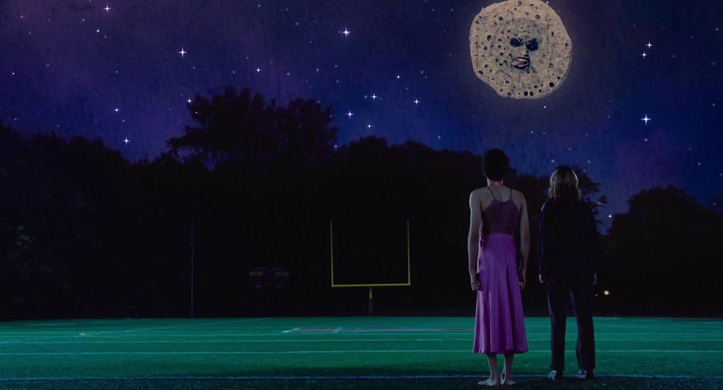 A wide shot of Owen (Justice Smith) and Maddy (Brigette Lundy-Paine) standing on a football field to the right of the frame looking at an artificial moon in the night sky with a grotesque expression on its face.