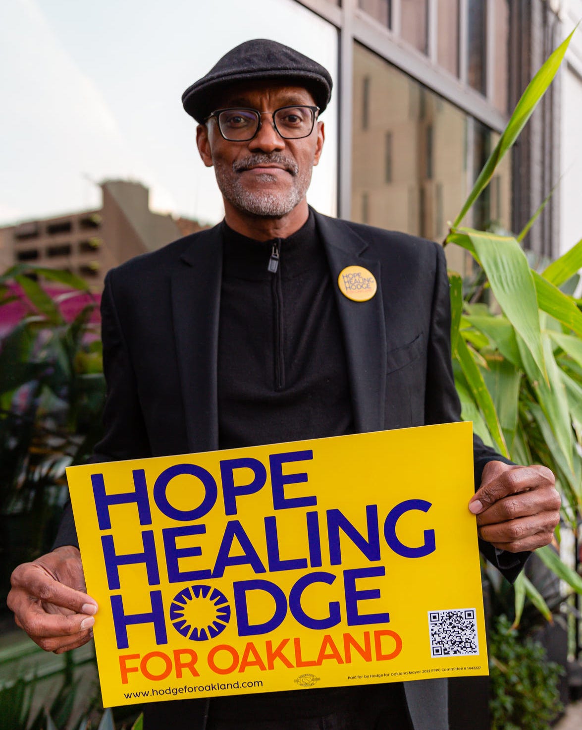 Greg standing, with a black cap and jacket, holding his campaign sign: "Hope, Healing, Hodge for Oakland"