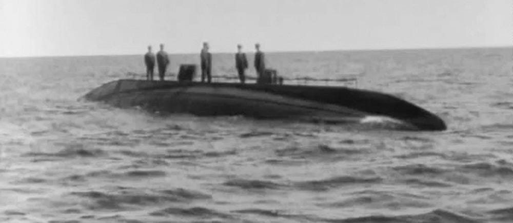 The submarine Nautilus, a life-sized vessel built for the film 20,000 Leagues Under the Sea in 1916, on a calm sea. On the top deck a group of five me stand around. 