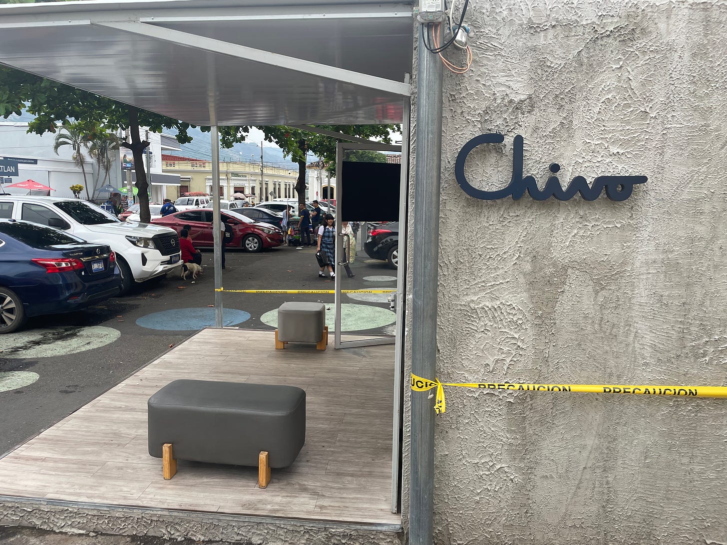 A concrete building surrounded by police tape sits in a parking lot, and the word "Chivo" is written in a sign in black letters on the wall