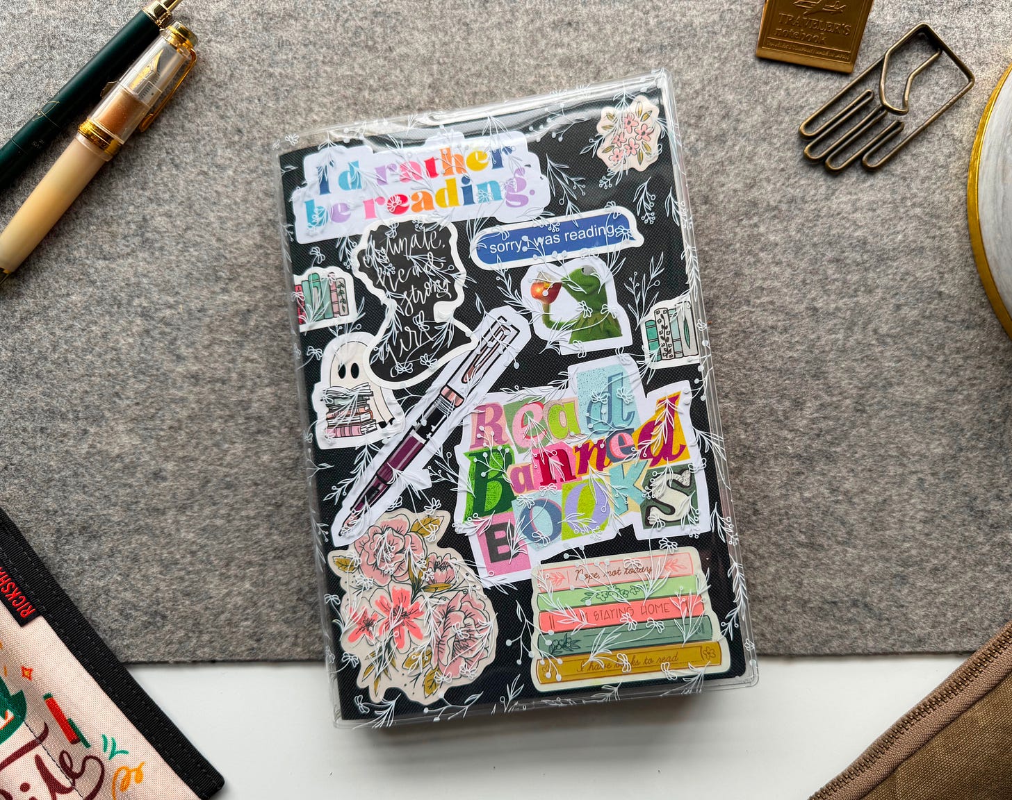 Overhead picture of an A5 notebook on a desk. A5 notebook is black and has various book related stickers on the cover, as well as a clear cover with a white floral print. Stationery items scattered around the notebook. 