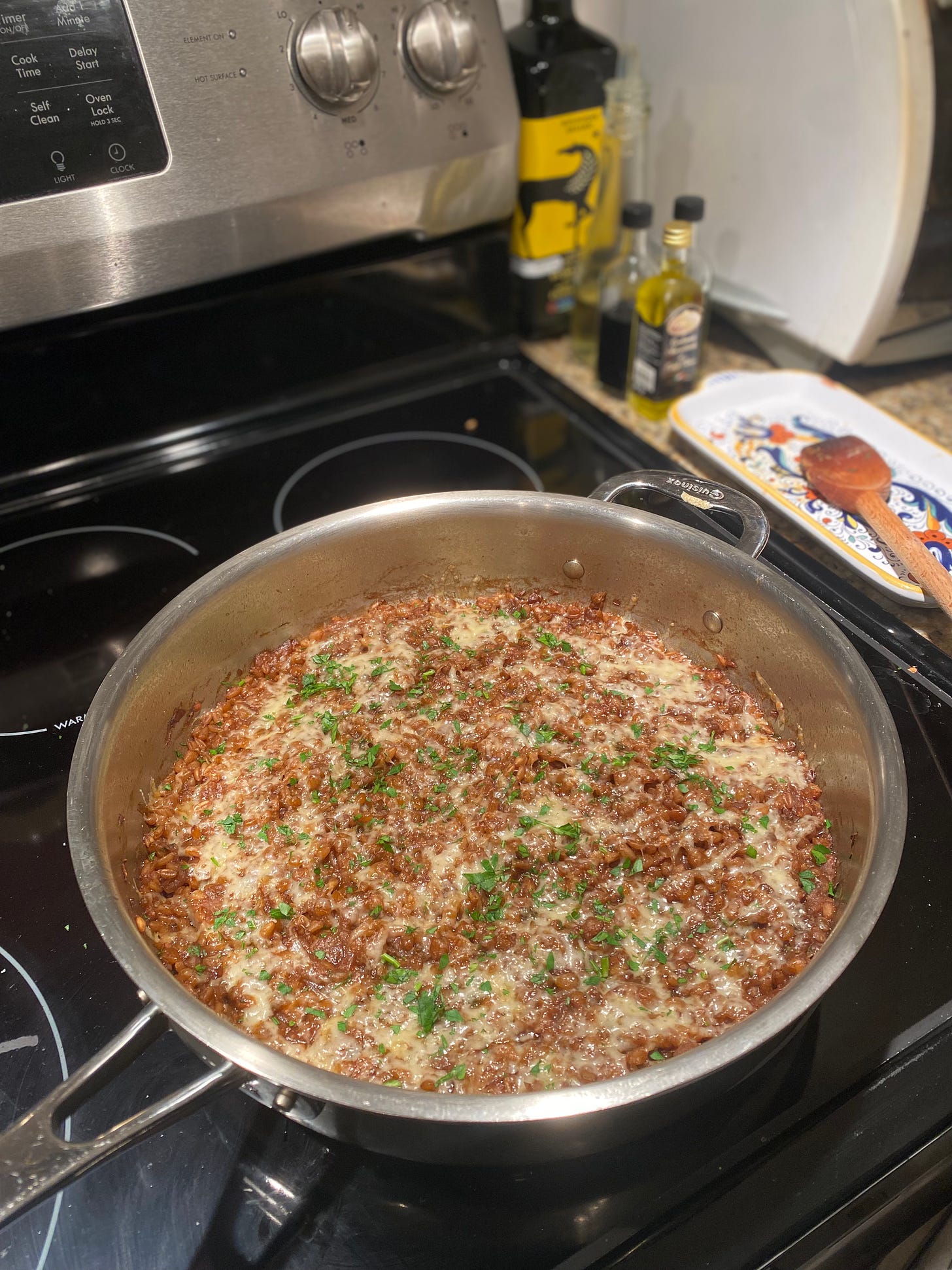 On top of the stove, a 12" steel pan full of the lentil farro stew described above. It has cheese melted over the top and sprinkles of parsley. Next to the stove is a spoon on its rest.