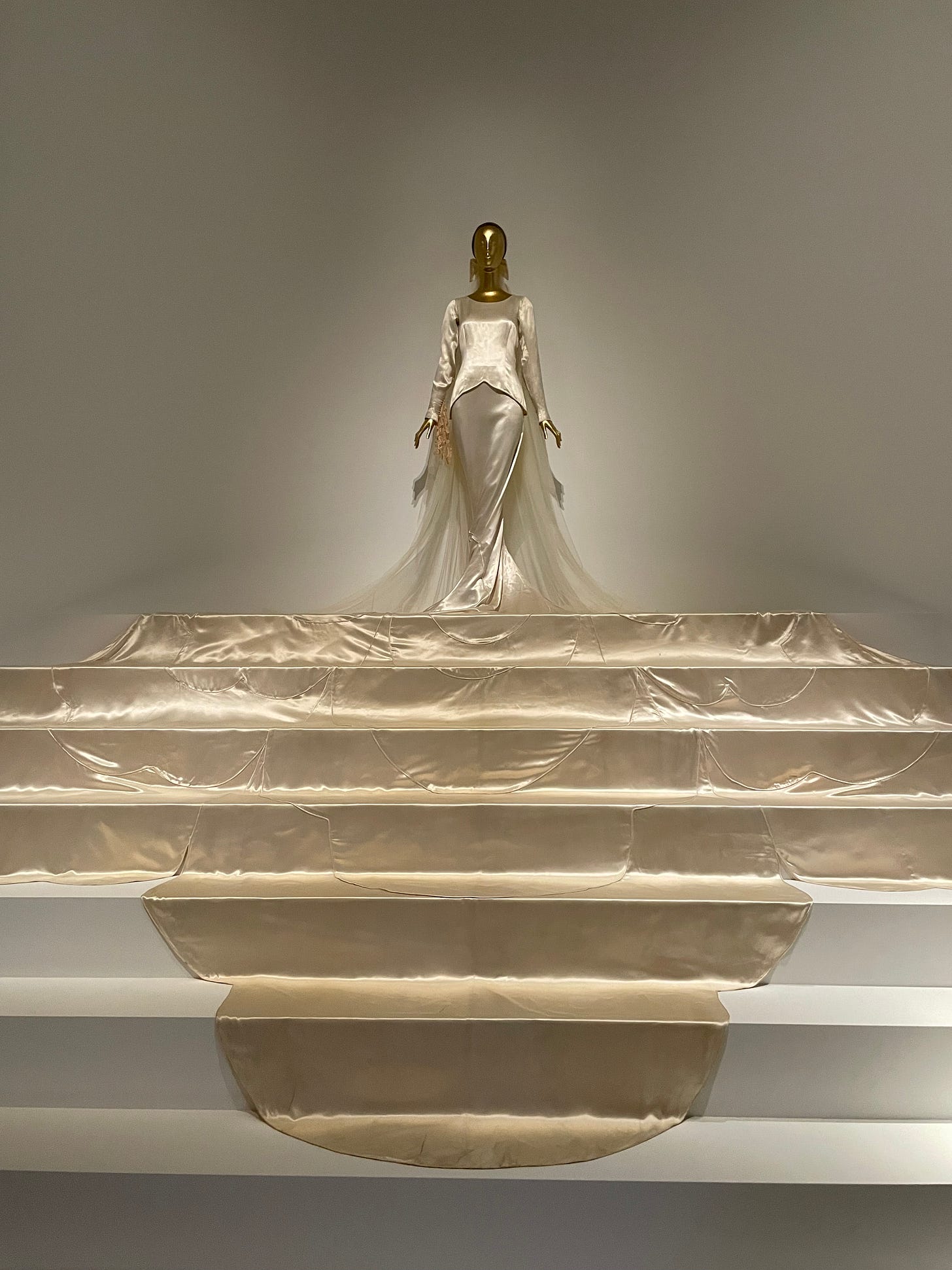 A metallic gold mannequin stands atop a white staircase wearing a cream-colored stain wedding dress with a dramatic train that cascades down multiple steps. 