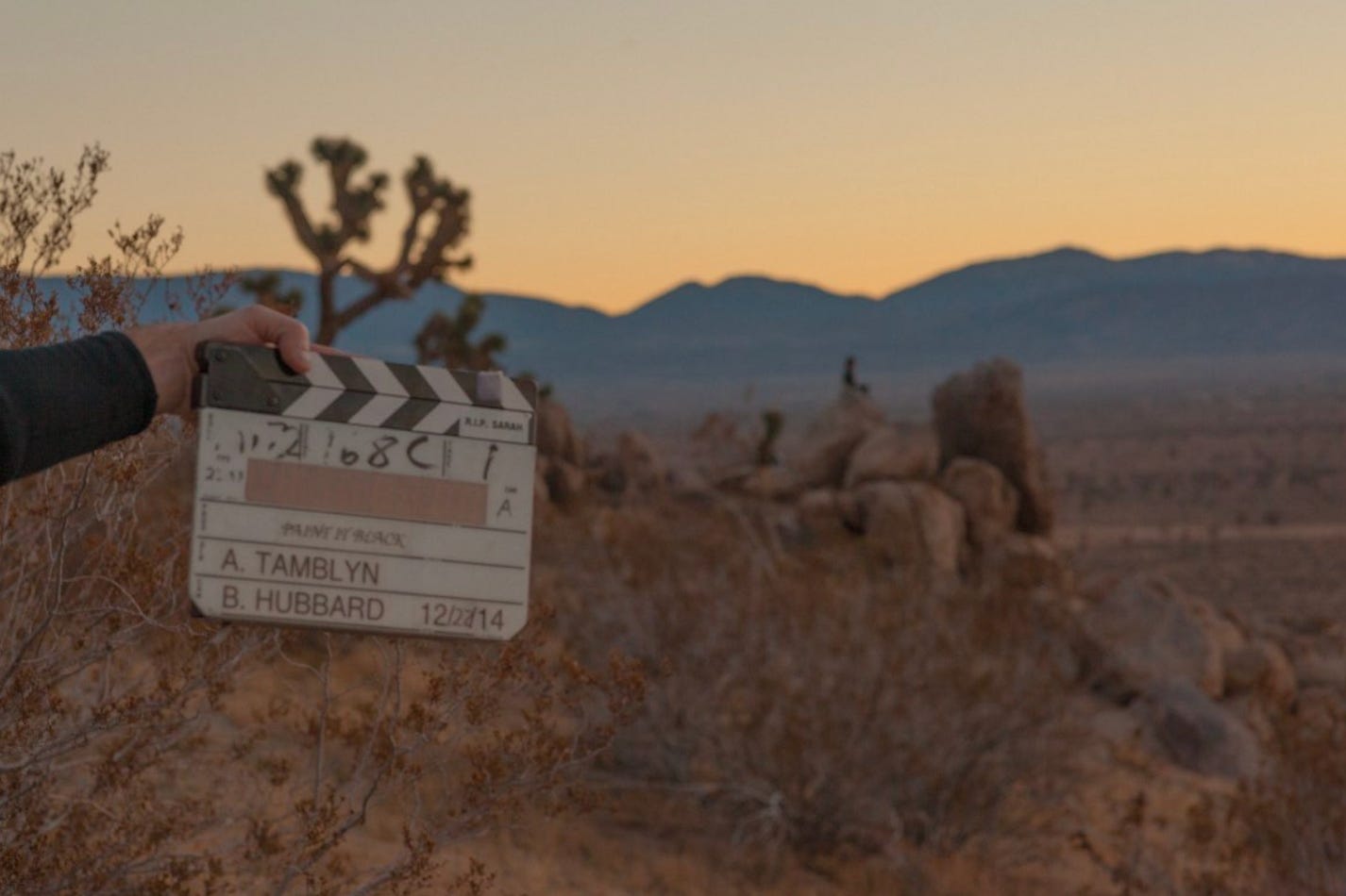 A hand holding a clapboard in the desert. The person's body is out of frame. The clapboard reads: Paint It Black A.Tamblyn / B. Hubbard / 12.22.14
