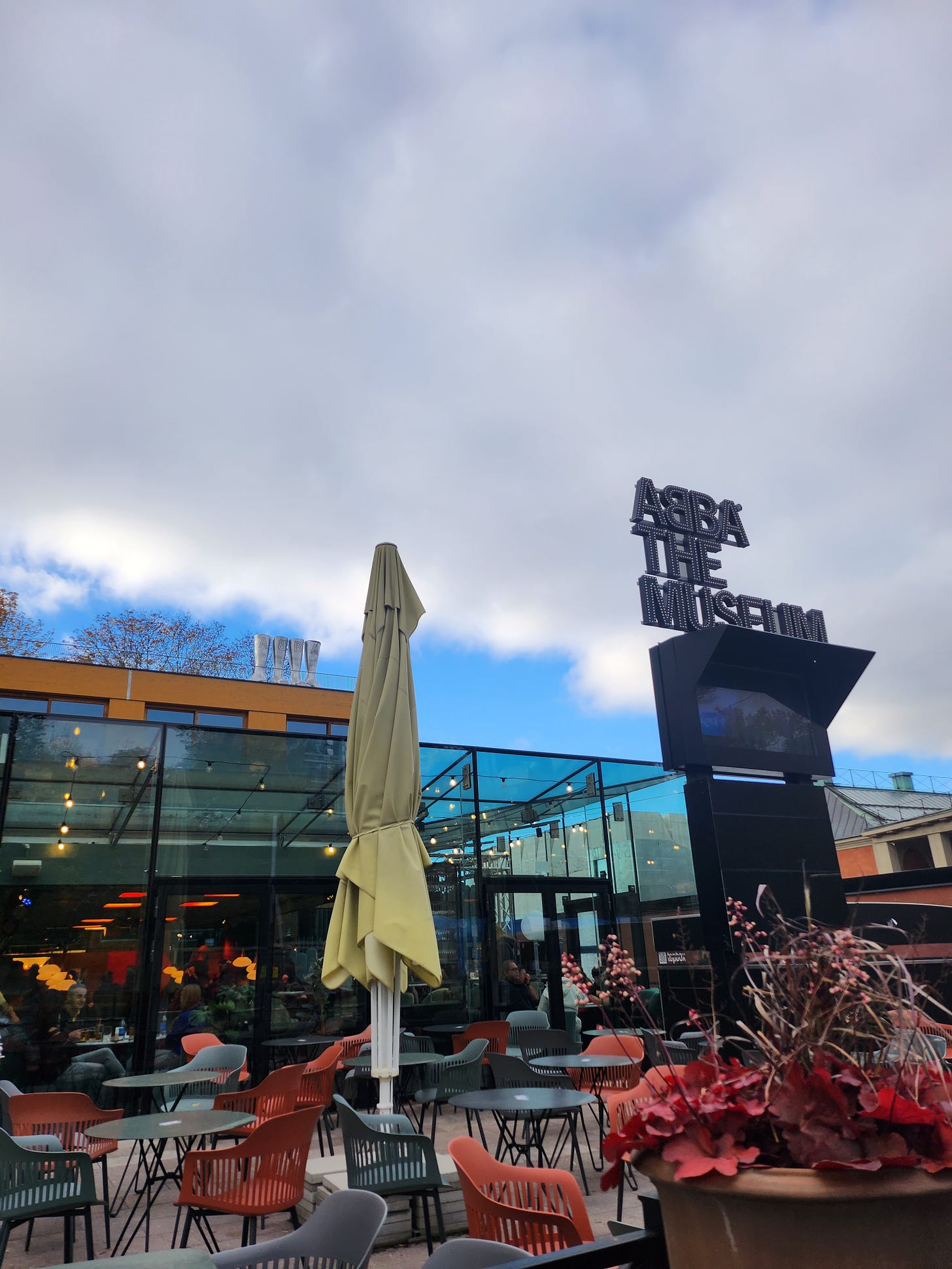 Against a cloudy sky, with just a hint of blue, the sign for Abba The Museum in black caps lock stands starkly. Below is an outdoor cafe setting with tables and chairs. 