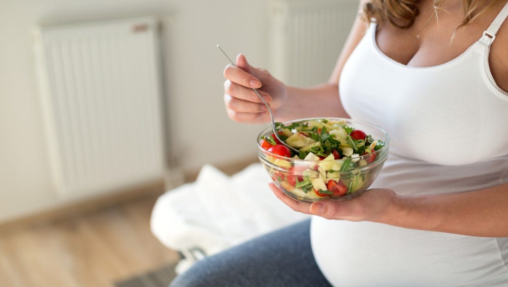 Pregnant woman sitting on edge of bed holding a bowl of veggies