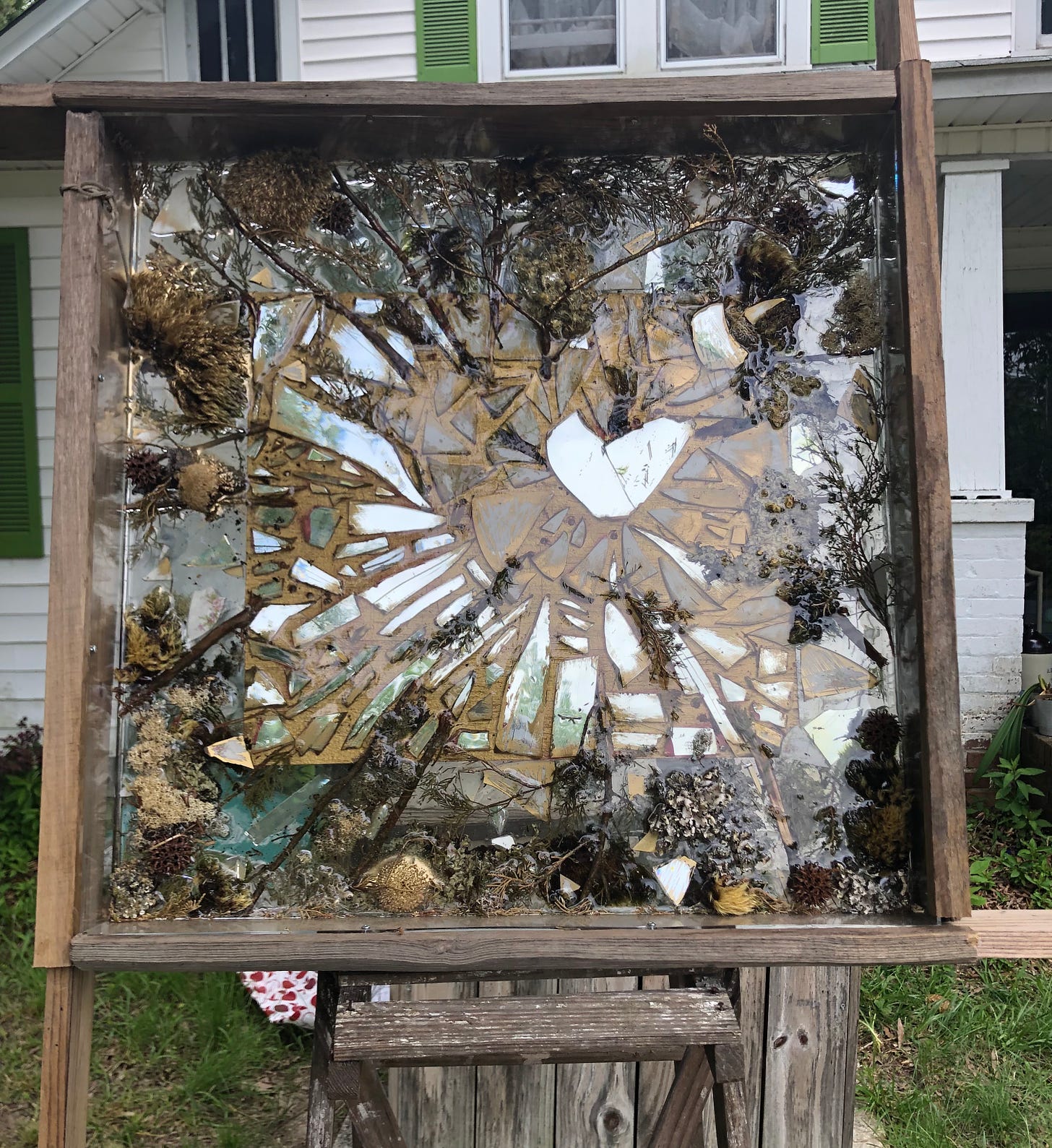 A mosaic made of glass with a heart at the center, surrounded by natural beach items, encased in a wooden box, sitting on a ladder, in front of a house.