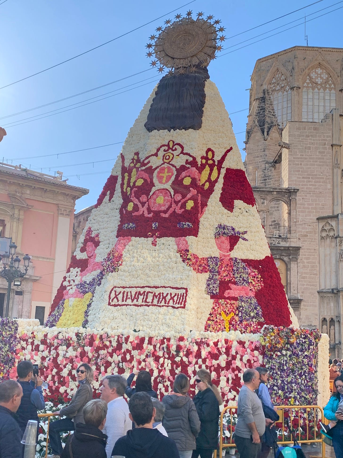The back of Mary's completed cape with an intricate crown being held up by a Fallero and Fallera, all in multicolored flowers.