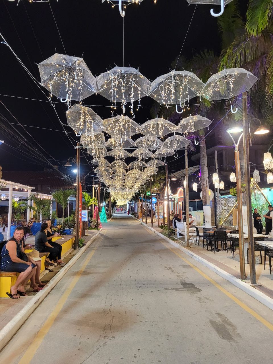 Uma rua estreita com sombrinhas transparentes penduradas, todas trazendo luzes de natal brancas penduradas em fios, o que as faz lembrar águas-vivas. Há pessoas sentadas em bancos nas laterais e dá pra ver que de ambos os lados tem estabelecimentos iluminados.