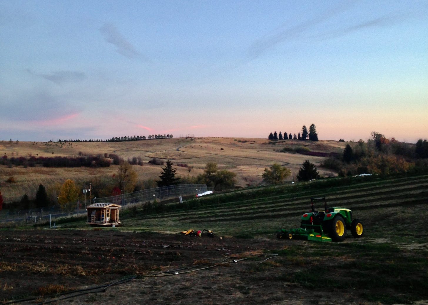 the organic farm in pullman, wa at sunset. a tractor in the right foreground, a small shack in the left, rolling hills in the background.