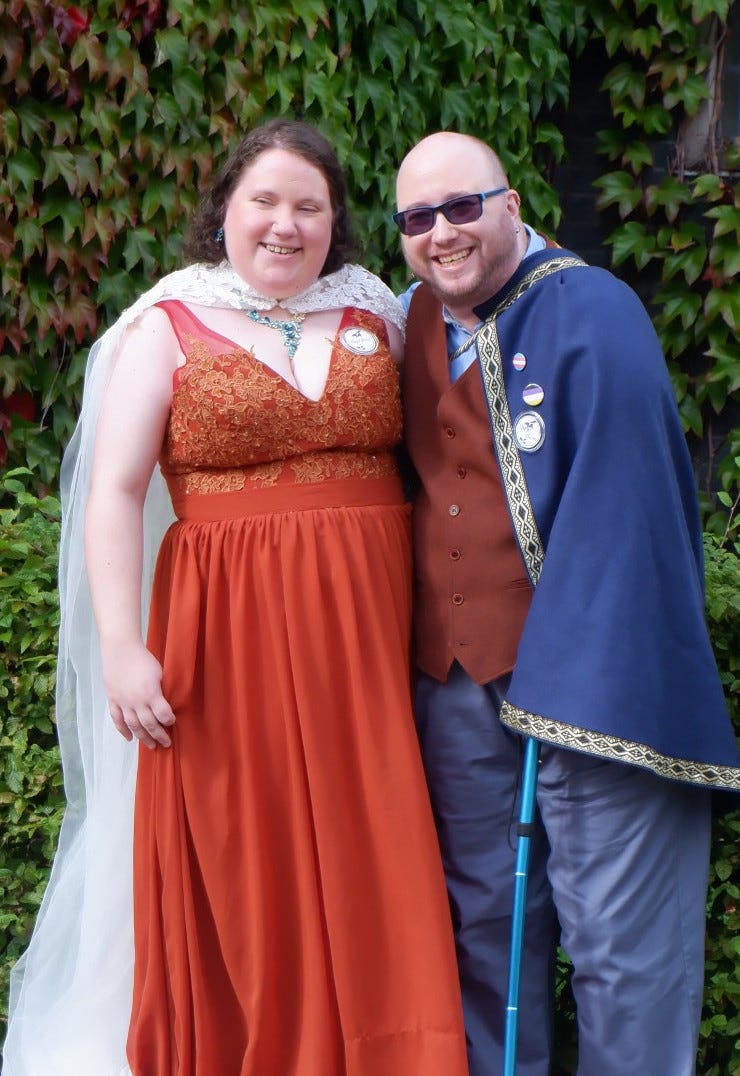 A woman with brown hair, curly at the bottom, who wears a burnt orange dress with embroidery, a blue necklace and matching dangly blur earring, and a white lacy train, stands next to a beardgender person, with a shaved head and close-trimmed facial hair, wearing black & blue glasses, a matching dangly blue earring, a burnt orange waistcoat, light blue shirt and trousers, and royal blue cape. Both are grinning towards the camera.