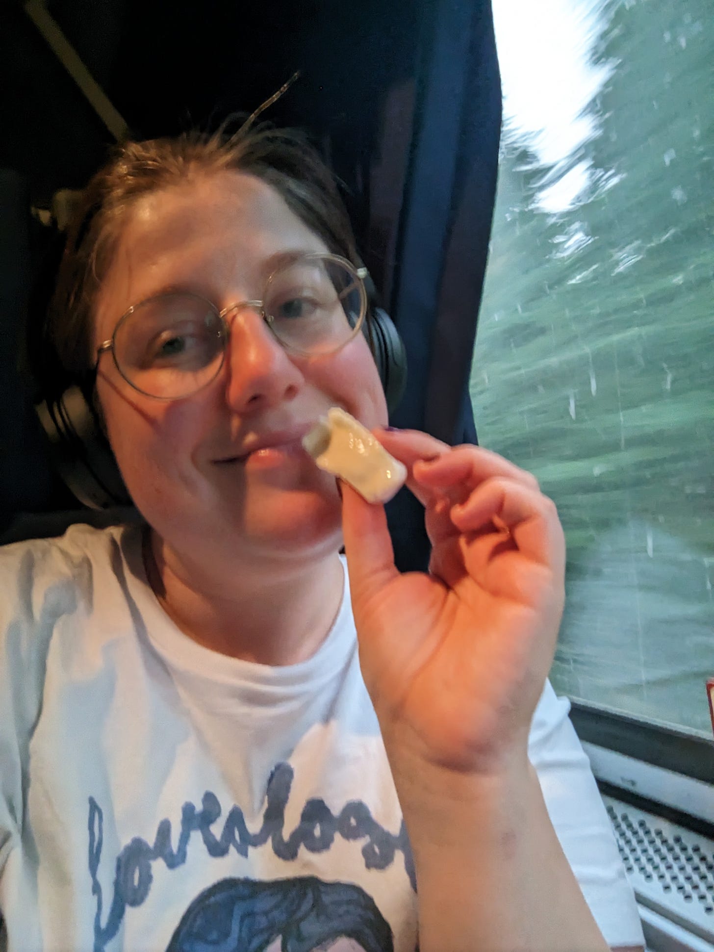 a white woman with brown hair pulled back and round glasses smiles and poses with a half-eated mini brie cheese. She is wearing over-ear headphones and a "loveology" t shirt and sitting next to a train window.