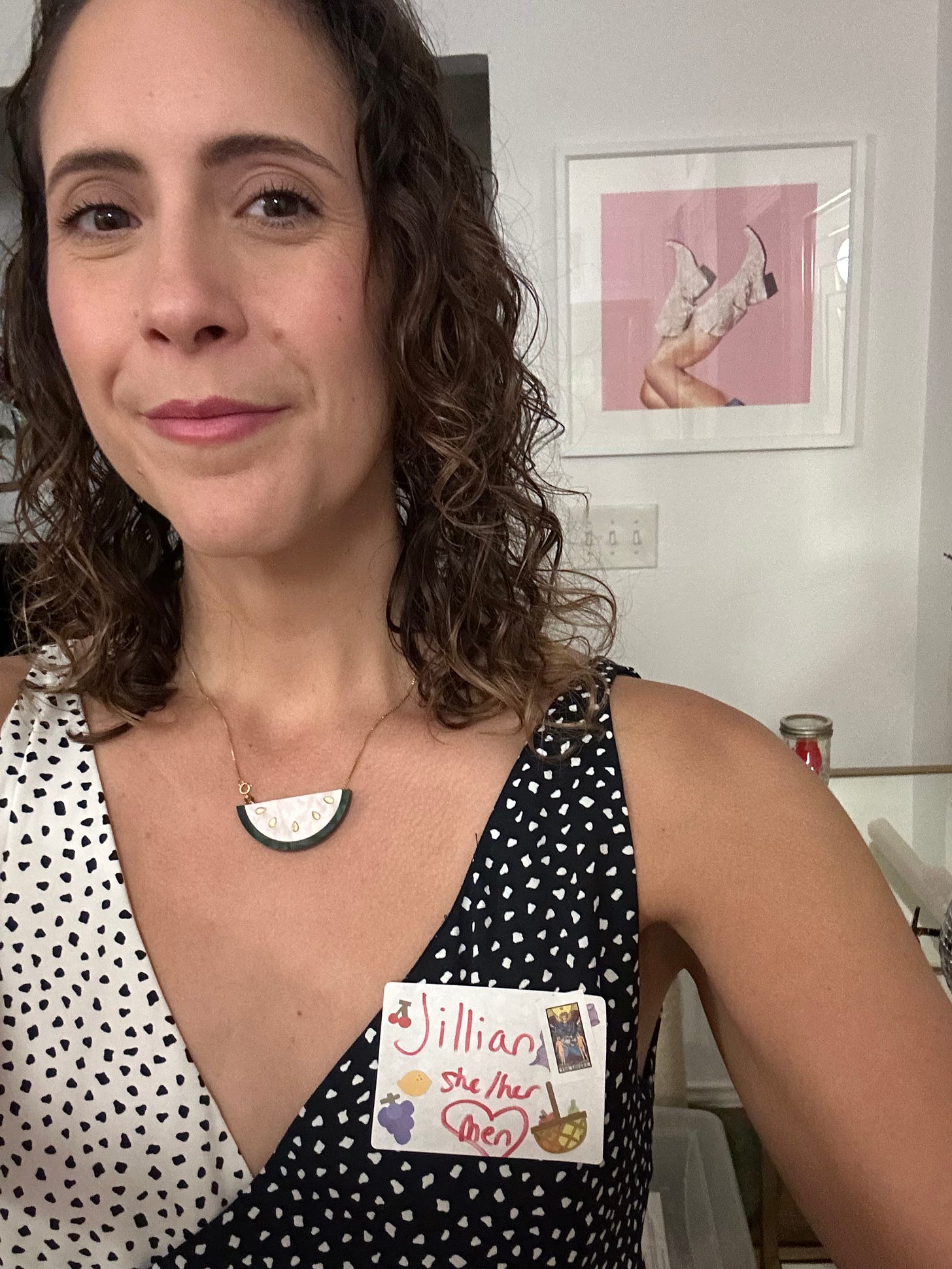 A brunette woman with curly hair smiles and takes a selfie. She wears a watermelon necklace, polka dot dress, and a nametag with her name, pronouns, and interest in men.