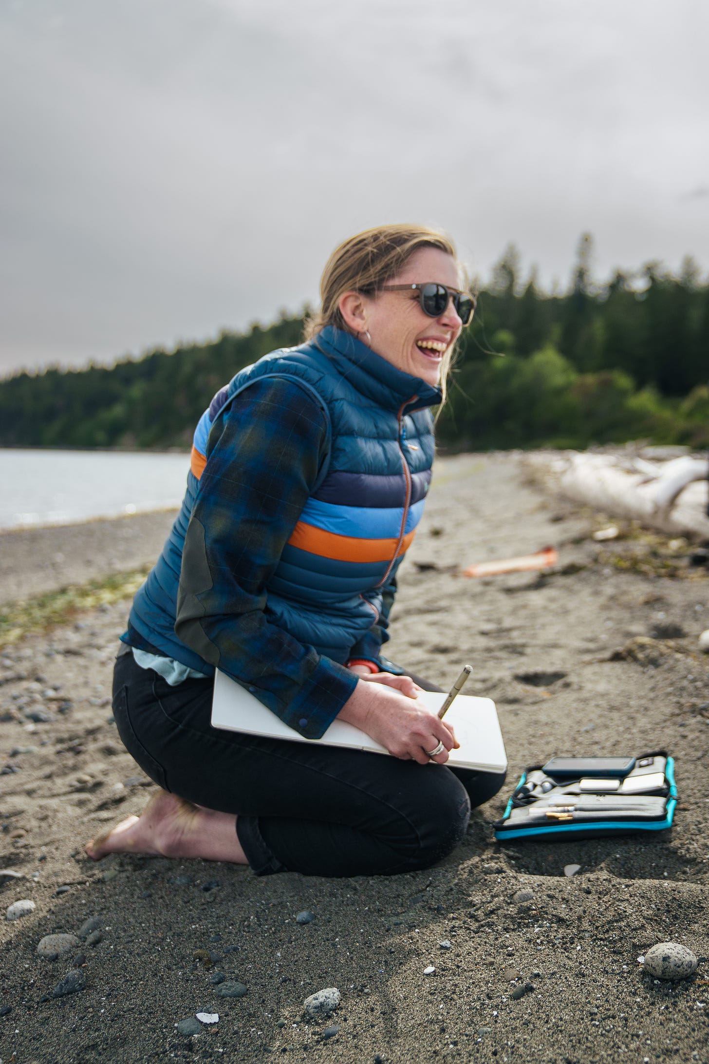 A photo of the artist Anna Brones, sitting on a sandy beach with trees and water behind her, and a notebook and pen in her hand, laughing and smiling 