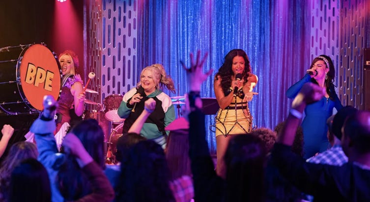 A screenshot from Girls5eva Season 3 featuring Summer, Gloria, Wickie, and Dawn performing on stage while a crowd cheers them on