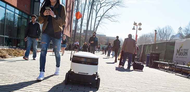 Press photo of a Starship delivery robot on a wide pavement surrounded by students.