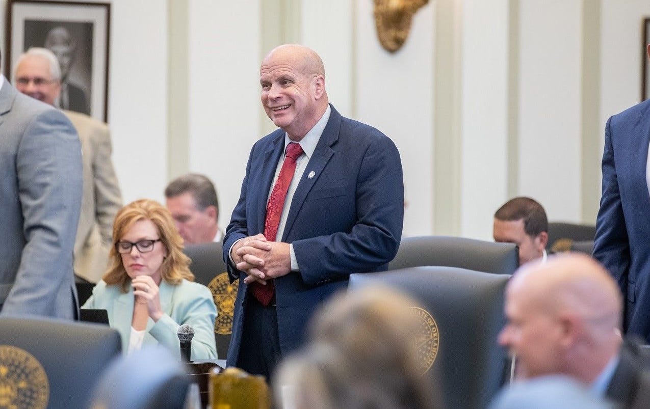 Jim Olsen, wearing a blue suit with red tie, stands among a small group of seated people.