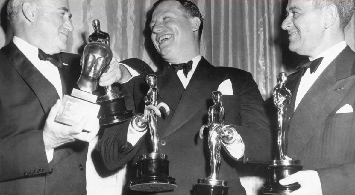 Harold Russel, usando smoking e sorrindo, segura duas estatuetas do Oscar com seus braços protéticos, que usam ganchos nas pontas. Ele é branco, tem cabelo escuro. A foto é em preto e branco. 