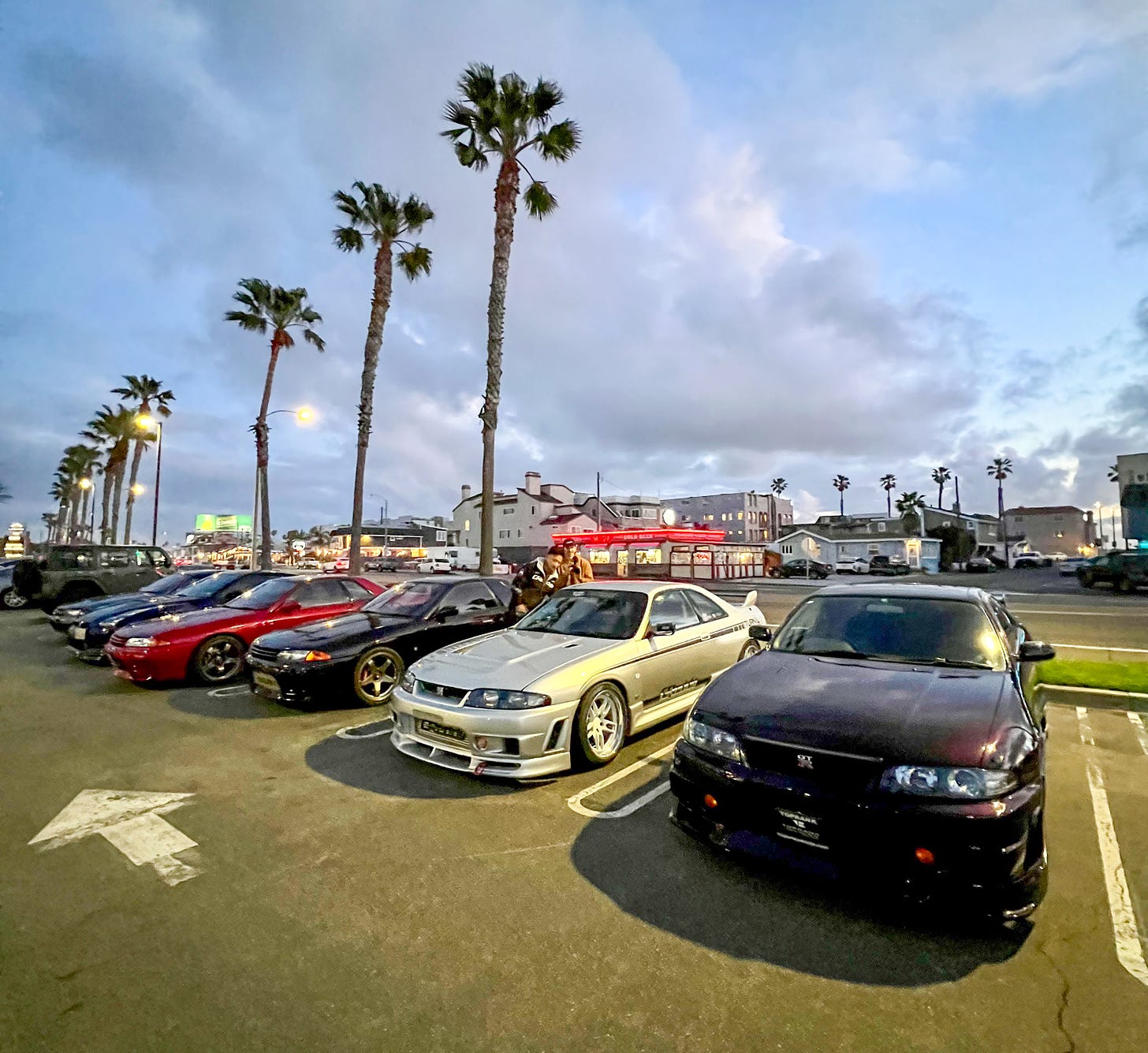 A half-dozen R32 and R33 Skyline GT-R sports cars take up a parking lot in Southern California during sunset with soft streetlights making them glow and palm trees towering overhead.