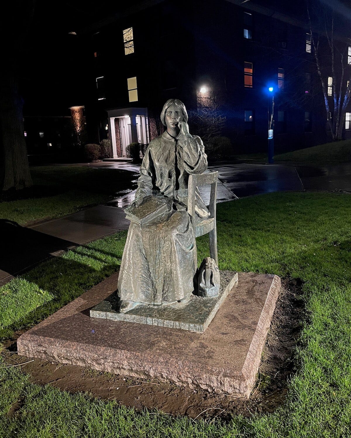 The Elizabeth Blackwell statue at HWS, at night. Elizabeth is sitting sideways on a wooden chair, book in her lap, and medical bag at her feet. Her elbow rests on the back of the chair, her chin on her her elbow, looking pensive.