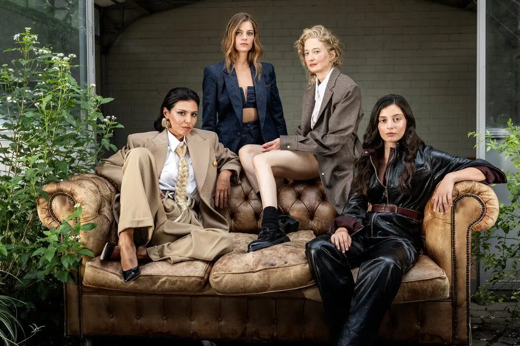 Two women sit on the seat of a leather sofa, another sits on its back and a fourth stands behind the sofa. All are wearing variations on suits, with quizzical expressions on their faces. 