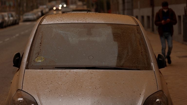 Photo of a parked car in a non-specific European city street, covered in fine red dust that has blown over from the Sahara desert.