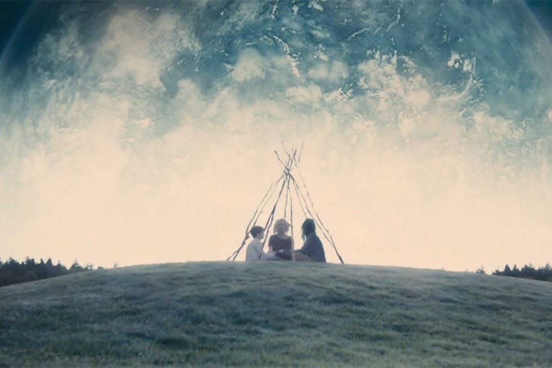 cena do filme “melancolia”, com três pessoas no alto de um monte sentadas embaixo de uma armação rústica de galhos, como se fosse uma cabaninha, vistas de longe, e ao fundo o planeta melancolia, azulado, gigantesco, se aproximando para acabar com a terra.