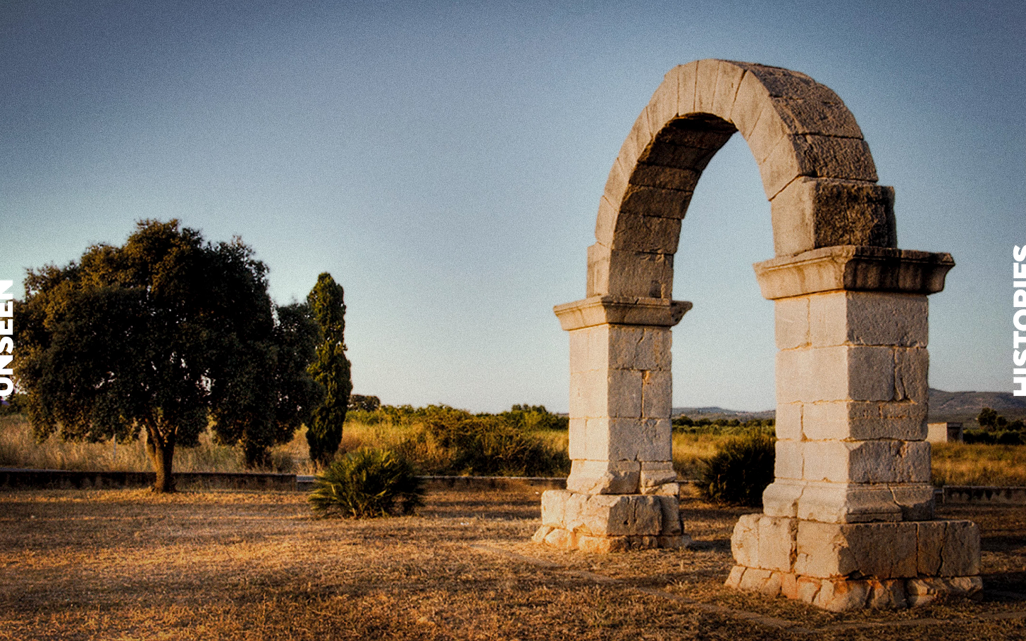 Colour photograph shows the stone Arch of Cabanes in Valencia. Brown grass and a blue sky.