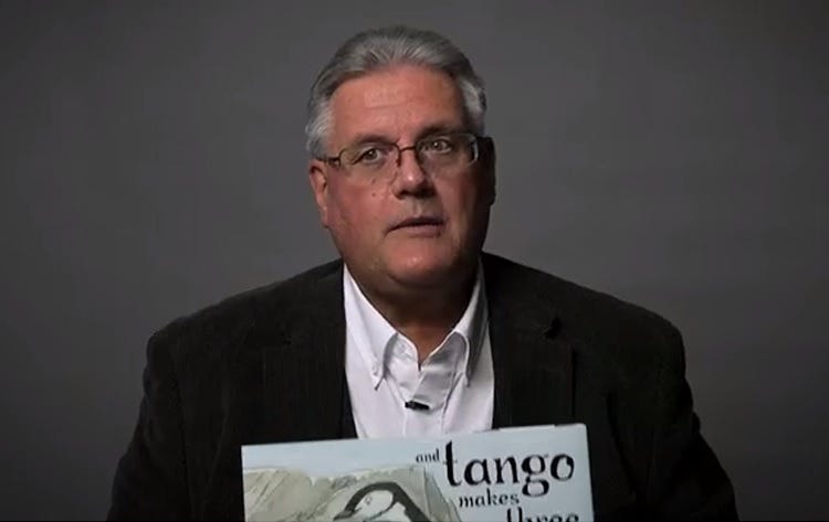 video screenshot of a white man with glasses and grey hair, holding a copy of 'And Tango Makes Three,' which he believes children lack the 'cognitive skill to process' because how can there be two daddy penguins???