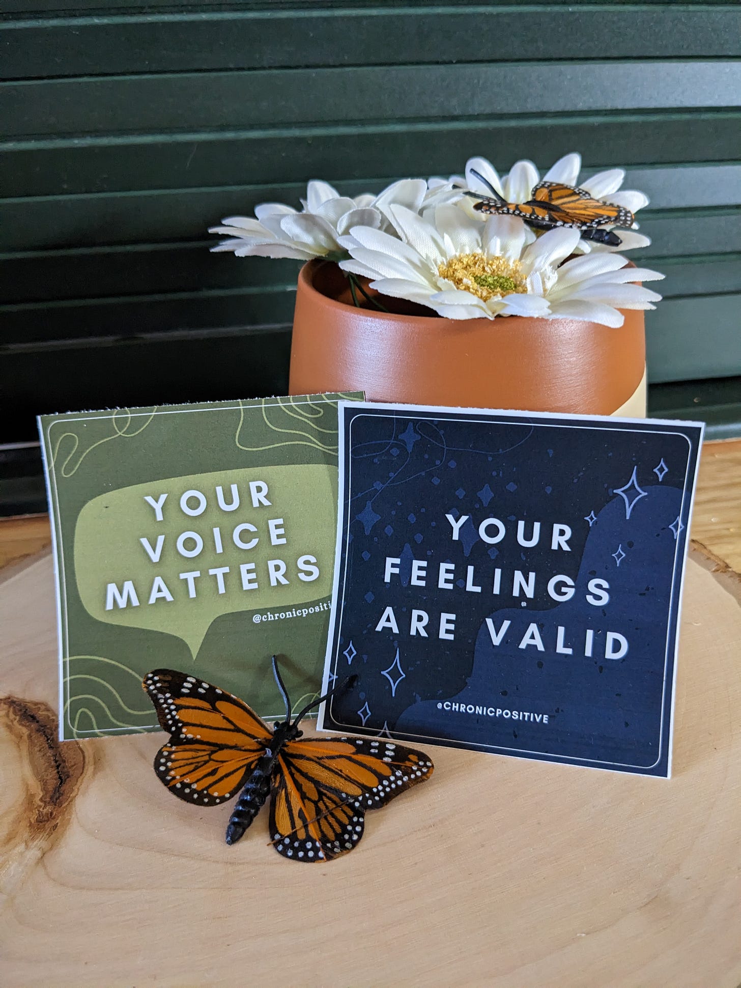 A photo of a green sticker that says "your voice matters," accented by a speech bubble, and a navy sticker that says "your feelings are valid," accented with sparkles. The stickers are staged with an earthy orange vase, daisies, and fake monarch butterflies.