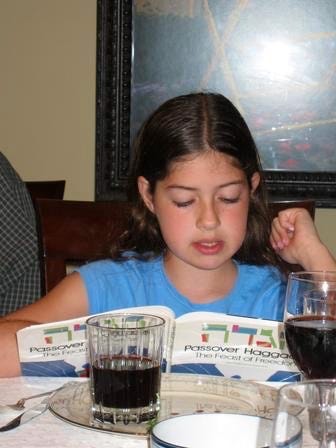 Young white femme child with long brown hair looking at a Passover Haggadah with a glass of wine on the table