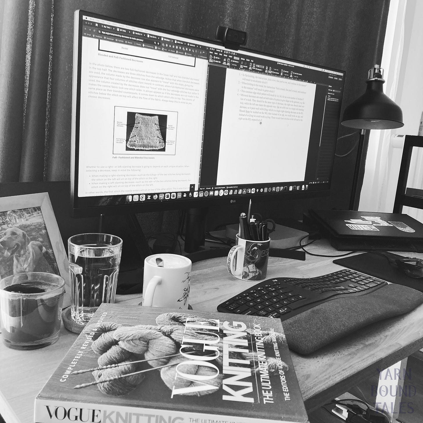 A black and white photo showing a workstation with a large monitor, a keyboard, a coffe mug, some water, the photo of a dog, a laptop, as well as the newest edition of Vogue Knitting, a knitting reference book.