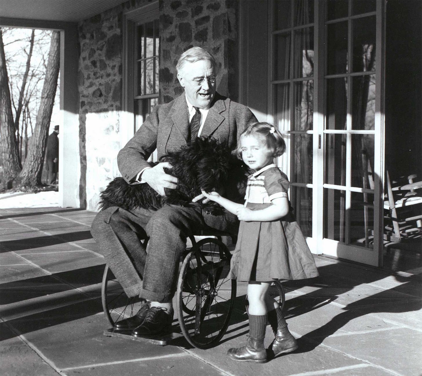President Roosevelt seated in a wheelchair with a puppy dog on his lap, next to a young girl.