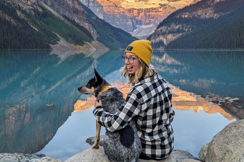 Scout, a blue heeler, and Haley, a young woman in a yellow hat, pose at Lake Louise in Banff National Park