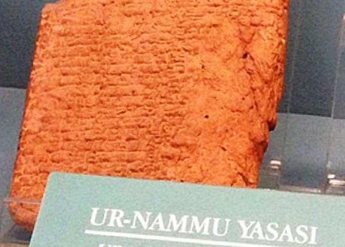 An upright slab of orange-colored clay imprinted with rows of tiny hatch marks. Slab is under glass in a museum, and the exhibit sign reads "Ur Nammu Yasasi," or Ur Nammu Law Code.
