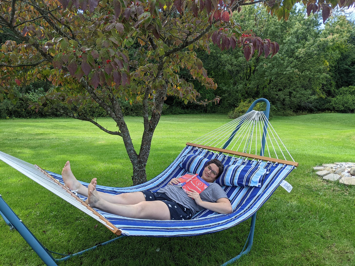 Photo of me lying on the hammock in the backyard. It's striped various shades of blue and sitting under a small tree.