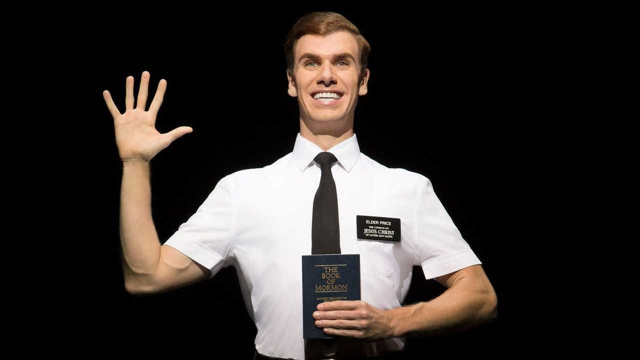 Promotional shot for The Book of Mormon with a smiling man holding up his hand to wave and holding a copy of the Book of Mormon in his other hand. He wears a short sleeved dress shirt and tie.