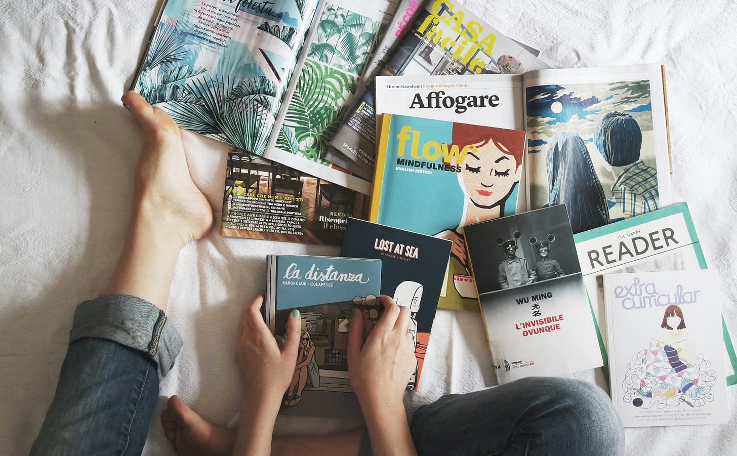 woman sitting on a bed in jeans with a collection of books and magazines scattered around her