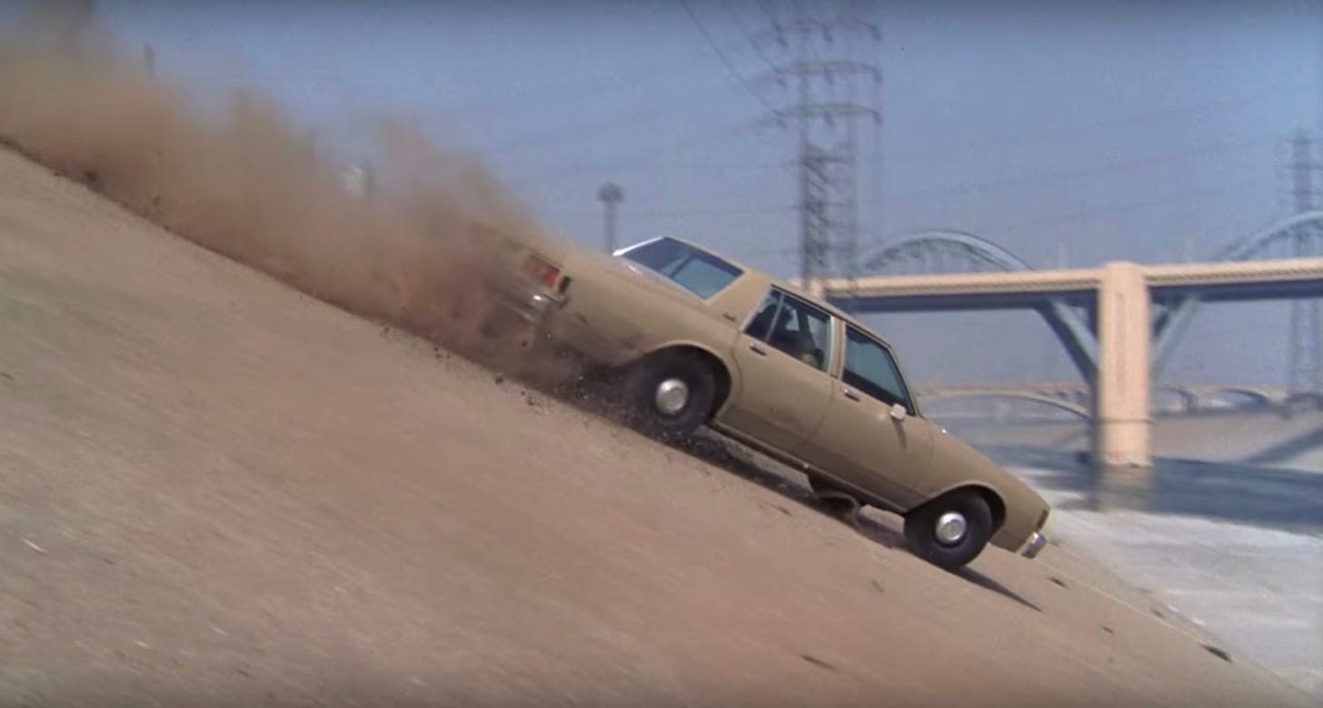 Still from a movie. An old crappy car drives down an embankment into the Los Angeles River