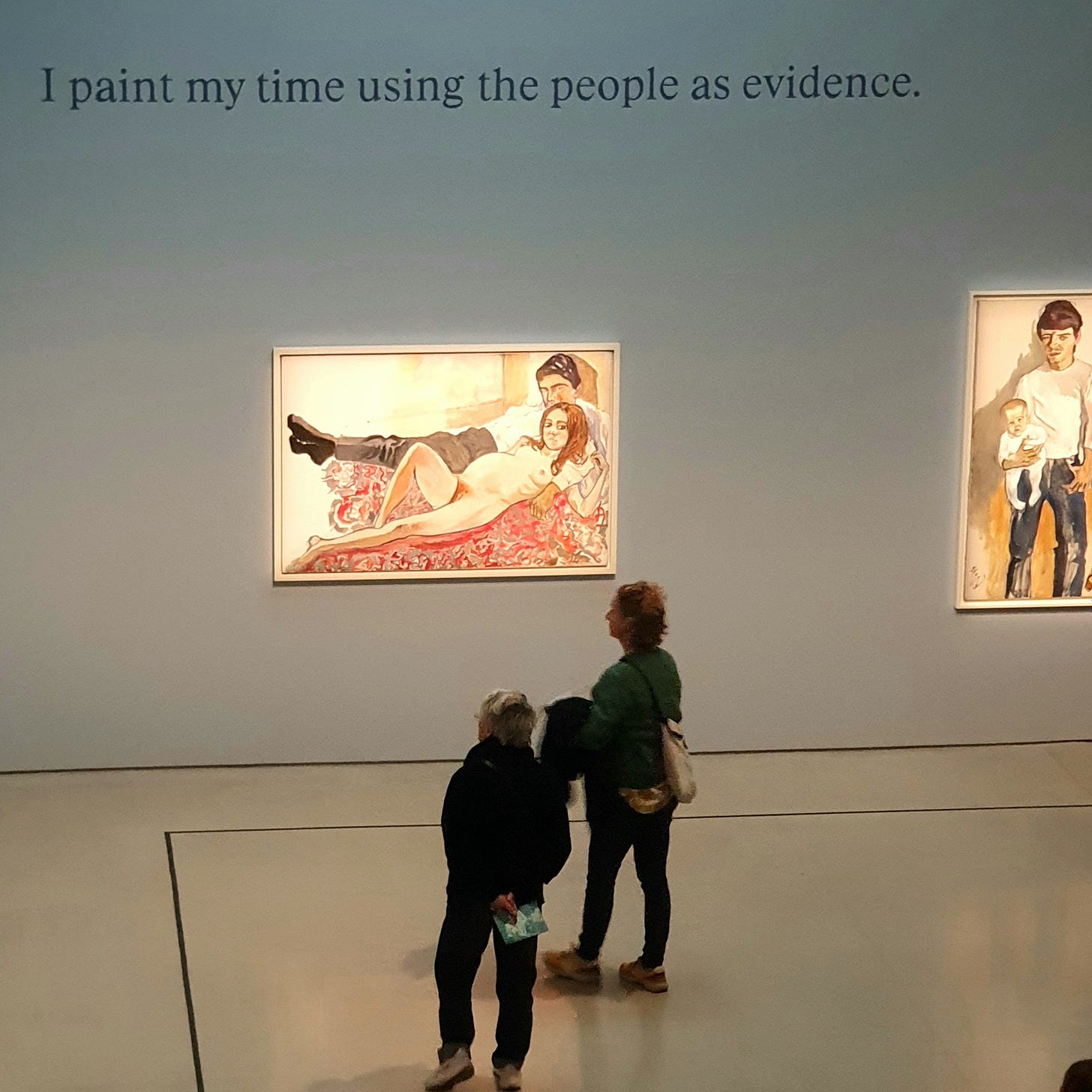 Photo of Alice Neel paintings and 2 onlookers. Caption on the gallery wall reads “I paint my time using the people as evidence”