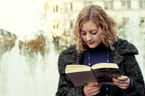 An old photo of Katie. She has dyed blonde hair, is wearing a black coat and is reading a copy of Atonement by Ian McEwan with a fountain just behind her.