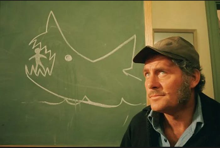 Quint from Jaws in front of a chalkboard drawing of a shark eating a guy (foreshadowing????)