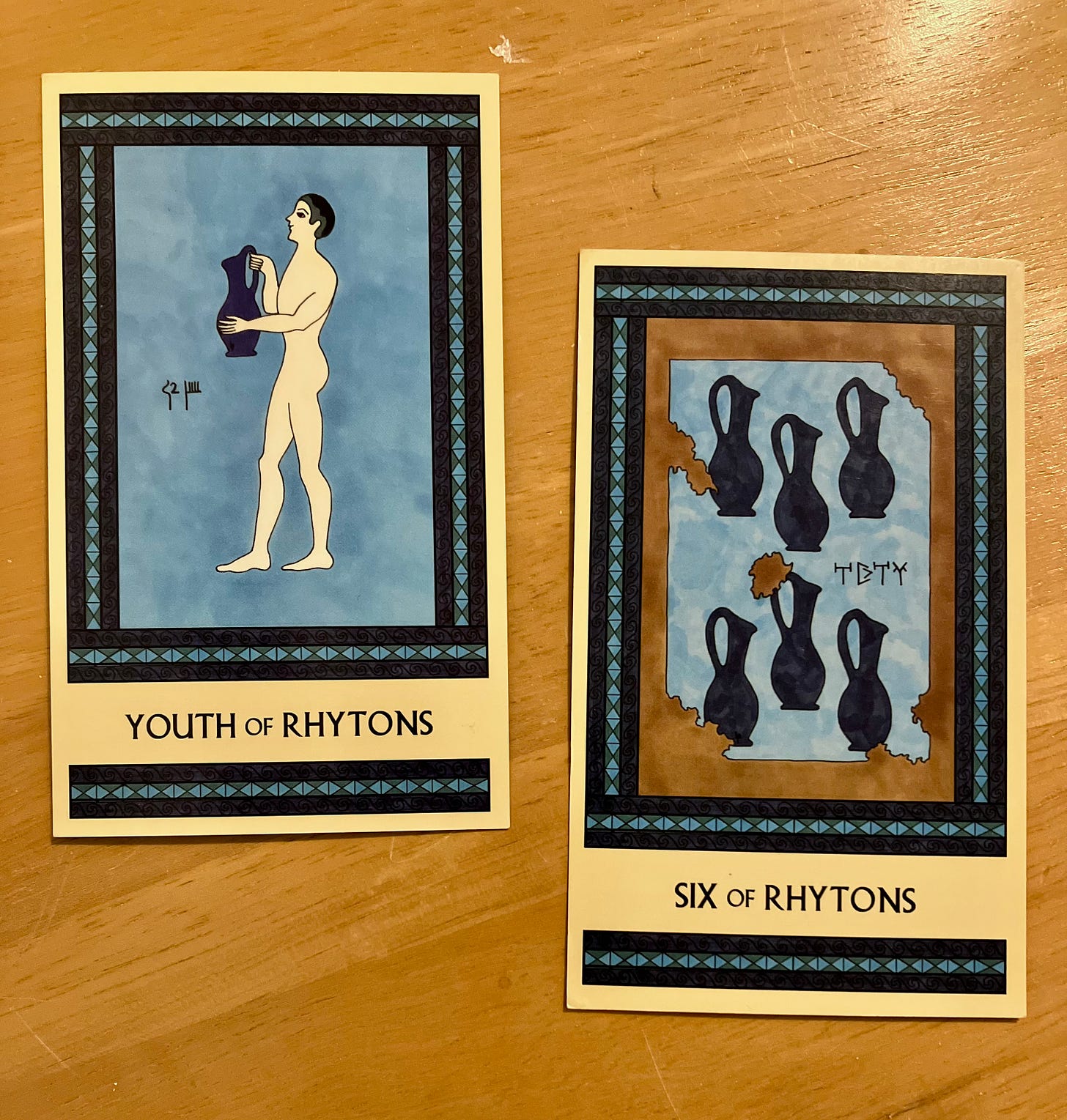 Two Minoan Tarot cards side by side on a golden wood surface. Both cards are painted in shades of blue. The Youth of Rhytons shows a boy standing, facing left, holding a large royal blue pitcher. The Six of Rhytons shows a crumbling fresco painting of six dark blue pitchers.