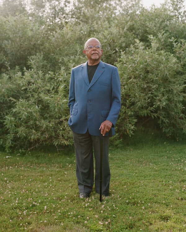 Henry Louis Gates Jr., wearing a blue suit jacket and gray trousers, stands in a patch of grass, one hand resting on a black cane. His chin is angled up as he looks into the far distance.