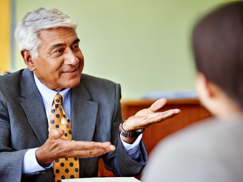 Profile of well-dressed brown-skinned man with gray hair dressed in blue suit and blue/yellow polka dot tie. He gestures with both hands to out-of-focus student to the right of the photo.