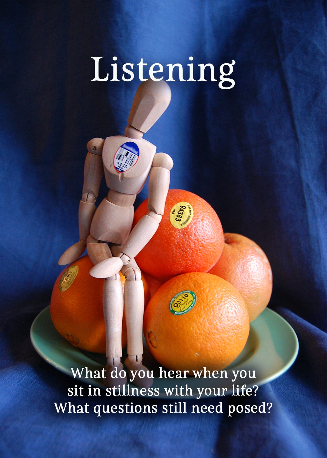 The Emotikin is sitting among a stack of oranges on a green plate, blue linen background, a quintessential still life portrait, only the manikin and oranges all have bar code labels from the grocery store. The image is titled Listening and the questions say What do you hear when you sit in stillness with your life? What questions still need posed?