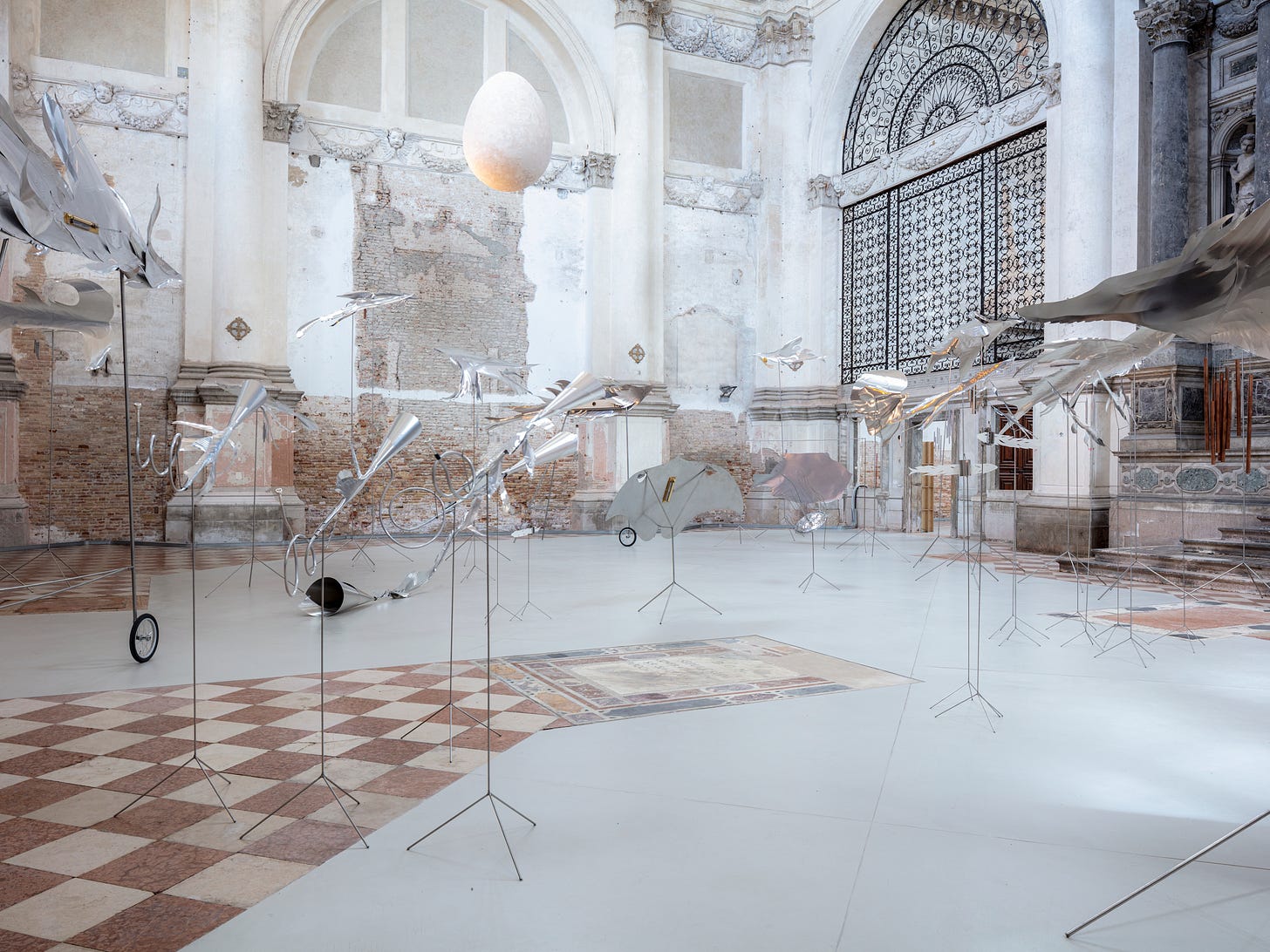 Inside a grand church with white, crumbly walls, aluminium objects and posts are arranged. The objects are shaped like sea animals in movement underneath an egg-shaped moon.